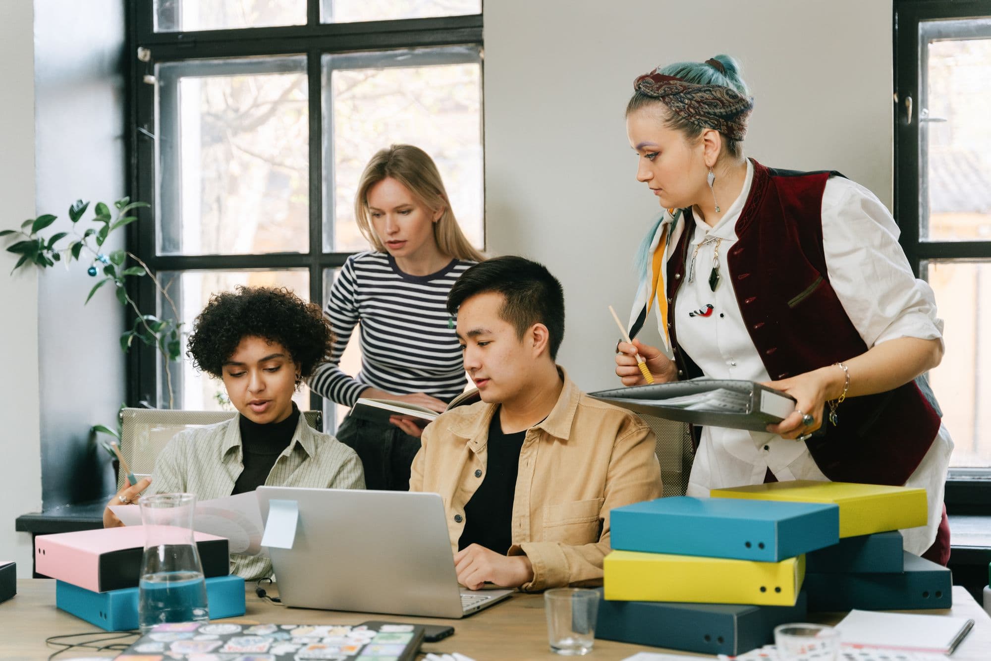 Four colleagues gathered around a laptop and a stack of colorful folders, reviewing paperwork together in a bright office